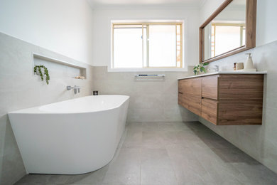 Design ideas for a mid-sized contemporary master bathroom in Hobart with medium wood cabinets, a freestanding tub, an open shower, a wall-mount toilet, beige tile, ceramic tile, yellow walls, ceramic floors, solid surface benchtops, grey floor, a hinged shower door, white benchtops, a niche, a single vanity and a freestanding vanity.
