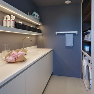 ロンドンの中サイズのコンテンポラリースタイルのおしゃれな家事室 (ダブルシンク、フラットパネル扉のキャビネット、白いキャビネット、人工大理石カウンター、青い壁、左右配置の洗濯機・乾燥機、ベージュの床、白いキッチンカウンター) の写真