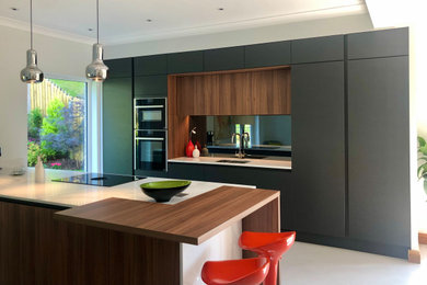Large contemporary grey and brown galley open plan kitchen in Glasgow with an integrated sink, flat-panel cabinets, medium wood cabinets, quartz worktops, metallic splashback, mirror splashback, black appliances, concrete flooring, a breakfast bar, grey floors and white worktops.