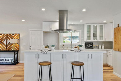 Small contemporary l-shaped open plan kitchen in San Francisco with a double-bowl sink, shaker cabinets, white cabinets, solid surface benchtops, white splashback, subway tile splashback, light hardwood floors, with island, beige floor and white benchtop.