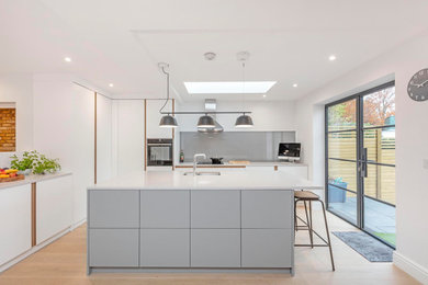Medium sized scandi grey and white l-shaped open plan kitchen in Surrey with a submerged sink, flat-panel cabinets, white cabinets, quartz worktops, grey splashback, glass sheet splashback, black appliances, light hardwood flooring, an island, beige floors, white worktops and feature lighting.