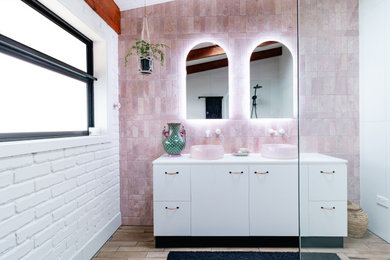 Photo of a mid-sized contemporary master bathroom in Melbourne with flat-panel cabinets, white cabinets, a curbless shower, a one-piece toilet, pink tile, stone tile, white walls, ceramic floors, a vessel sink, engineered quartz benchtops, brown floor, an open shower, white benchtops, a double vanity, a freestanding vanity, exposed beam and brick walls.
