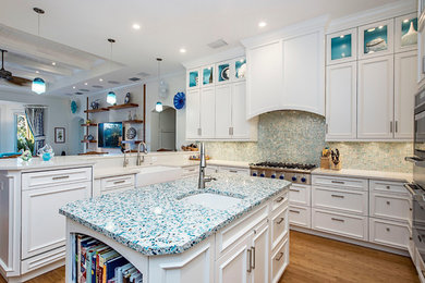 Example of a large beach style u-shaped medium tone wood floor and brown floor open concept kitchen design in Miami with a farmhouse sink, shaker cabinets, white cabinets, recycled glass countertops, blue backsplash, mosaic tile backsplash, stainless steel appliances and an island