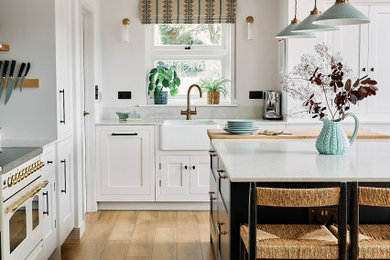 Medium sized rural l-shaped kitchen/diner in Berkshire with a belfast sink, shaker cabinets, white cabinets, engineered stone countertops, blue splashback, ceramic splashback, integrated appliances, medium hardwood flooring, an island, brown floors, white worktops, a vaulted ceiling and feature lighting.