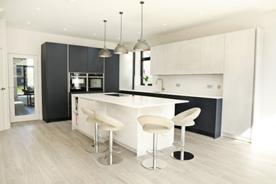 Photo of an expansive contemporary l-shaped kitchen/diner in London with a built-in sink, flat-panel cabinets, blue cabinets, quartz worktops, white splashback, stone slab splashback, black appliances, laminate floors, an island, grey floors, white worktops and feature lighting.