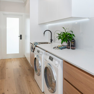 Modelo de cuarto de lavado de galera, actual, grande, con fregadero bajoencimera, puertas de armario de madera clara, encimera de cuarzo compacto, paredes blancas, suelo de madera clara, lavadora y secadora juntas y encimeras blancas