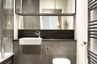 This is an example of a small modern master bathroom in London with glass-front cabinets, dark wood cabinets, a wall-mount toilet, ceramic floors, laminate benchtops and grey floor.