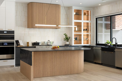 Trendy l-shaped light wood floor and beige floor kitchen photo in Vancouver with a farmhouse sink, gray cabinets, beige backsplash, stainless steel appliances, an island, black countertops and flat-panel cabinets
