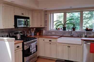 Eat-in kitchen - large cottage l-shaped medium tone wood floor eat-in kitchen idea in Milwaukee with a farmhouse sink, shaker cabinets, distressed cabinets, wood countertops, gray backsplash, stone tile backsplash, stainless steel appliances and no island