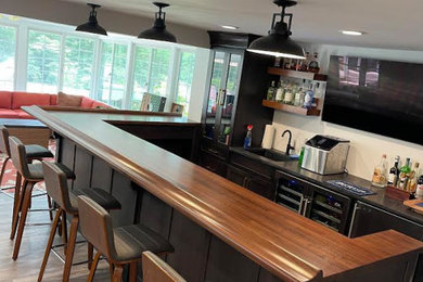 This is an example of a large transitional l-shaped seated home bar in New York with an undermount sink, shaker cabinets, dark wood cabinets, wood benchtops, vinyl floors, beige floor and brown benchtop.