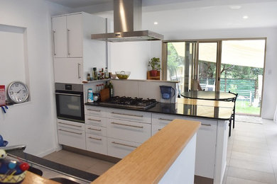 Photo of a small contemporary galley eat-in kitchen in Gold Coast - Tweed with white cabinets, granite benchtops, ceramic floors, with island, grey floor, black benchtop, a drop-in sink, open cabinets, white splashback, timber splashback and black appliances.