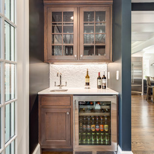 ワシントンD.C.の小さいトランジショナルスタイルのおしゃれなウェットバー (アンダーカウンターシンク、ガラス扉のキャビネット、中間色木目調キャビネット、クオーツストーンカウンター、白いキッチンパネル、石タイルのキッチンパネル、無垢フローリング、茶色い床、白いキッチンカウンター) の写真