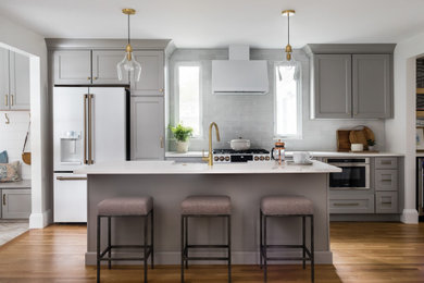 Design ideas for a medium sized classic single-wall kitchen/diner in Boston with a belfast sink, recessed-panel cabinets, grey cabinets, engineered stone countertops, white splashback, glass tiled splashback, white appliances, dark hardwood flooring, an island, brown floors and white worktops.