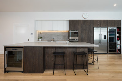 Photo of a large contemporary single-wall eat-in kitchen in Auckland with with island, a double-bowl sink, flat-panel cabinets, dark wood cabinets, solid surface benchtops, grey splashback, stone slab splashback, stainless steel appliances, light hardwood floors, grey benchtop and beige floor.
