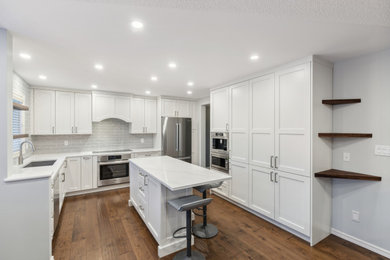Inspiration for a contemporary medium tone wood floor and brown floor kitchen remodel in Edmonton with a double-bowl sink, recessed-panel cabinets, white cabinets, quartz countertops, white backsplash, ceramic backsplash, stainless steel appliances, an island and white countertops