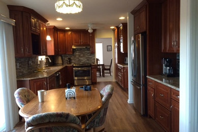 Photo of a mid-sized traditional l-shaped eat-in kitchen in Philadelphia with an undermount sink, raised-panel cabinets, medium wood cabinets, granite benchtops, beige splashback, stone tile splashback, stainless steel appliances, light hardwood floors, no island and beige floor.