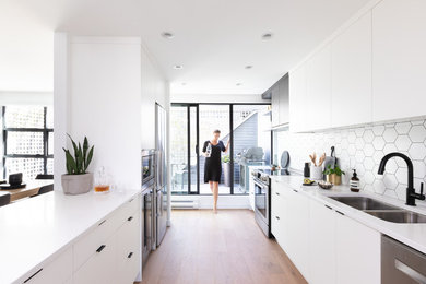 Trendy galley medium tone wood floor and brown floor kitchen photo in Vancouver with a double-bowl sink, flat-panel cabinets, white cabinets, white backsplash, stainless steel appliances, a peninsula and white countertops