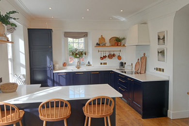 Large traditional u-shaped open plan kitchen in Cornwall with a submerged sink, recessed-panel cabinets, blue cabinets, quartz worktops, glass sheet splashback, integrated appliances, medium hardwood flooring and a breakfast bar.
