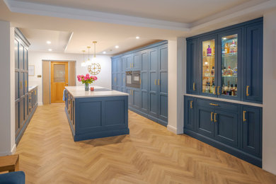 Photo of an expansive classic galley open plan kitchen in Manchester with a built-in sink, shaker cabinets, blue cabinets, quartz worktops, black appliances, medium hardwood flooring, an island, white worktops, exposed beams and feature lighting.
