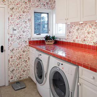 オマハのトラディショナルスタイルのおしゃれな洗濯室 (レイズドパネル扉のキャビネット、白いキャビネット、タイルカウンター、マルチカラーの壁、左右配置の洗濯機・乾燥機、グレーの床、赤いキッチンカウンター) の写真