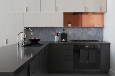 This is an example of a contemporary l-shaped kitchen in New York with an undermount sink, flat-panel cabinets, grey cabinets, grey splashback, stone slab splashback, black appliances, a peninsula, grey floor and black benchtop.