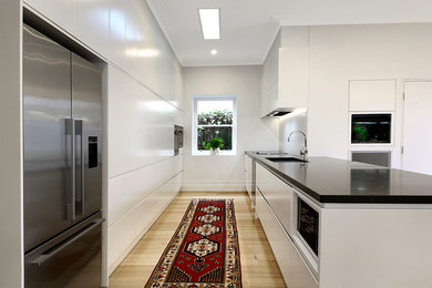 Large contemporary galley eat-in kitchen in Melbourne with an undermount sink, white cabinets, quartz benchtops, white splashback, stainless steel appliances, light hardwood floors, flat-panel cabinets, stone tile splashback and with island.