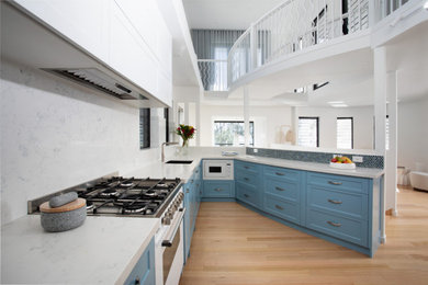 Photo of a medium sized nautical galley kitchen pantry with a submerged sink, shaker cabinets, blue cabinets, engineered stone countertops, multi-coloured splashback, mosaic tiled splashback, white appliances, light hardwood flooring, an island, brown floors and white worktops.