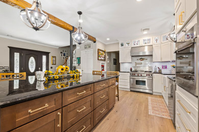Example of a large transitional l-shaped medium tone wood floor, brown floor and exposed beam eat-in kitchen design in Newark with a farmhouse sink, shaker cabinets, white cabinets, granite countertops, white backsplash, subway tile backsplash, stainless steel appliances, an island and black countertops