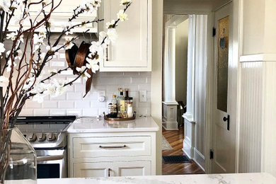 Mid-sized cottage l-shaped laminate floor and brown floor enclosed kitchen photo in Cedar Rapids with a farmhouse sink, beaded inset cabinets, white cabinets, quartz countertops, white backsplash, porcelain backsplash, stainless steel appliances, an island and white countertops