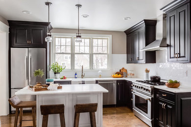 Elegant l-shaped medium tone wood floor and brown floor kitchen photo in Boston with a farmhouse sink, raised-panel cabinets, white backsplash, ceramic backsplash, stainless steel appliances, an island and black cabinets
