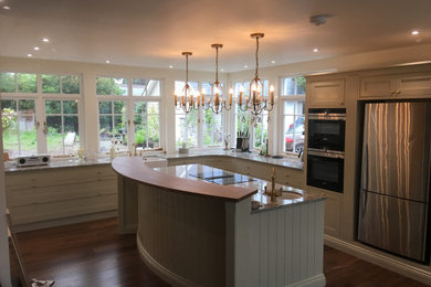 This is an example of a large contemporary kitchen/diner in West Midlands with a belfast sink, shaker cabinets, beige cabinets, granite worktops, stainless steel appliances, medium hardwood flooring, an island, brown floors and beige worktops.