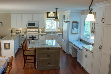 Large country u-shaped open plan kitchen in Toronto with a farmhouse sink, recessed-panel cabinets, white cabinets, granite benchtops, grey splashback, ceramic splashback, white appliances, medium hardwood floors, with island, brown floor and multi-coloured benchtop.