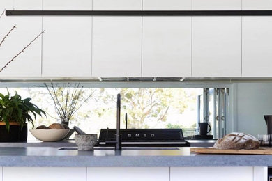 Mid-sized modern single-wall open plan kitchen in Sydney with a double-bowl sink, white cabinets, concrete benchtops, metallic splashback, mirror splashback, black appliances, with island, grey floor, grey benchtop and ceramic floors.