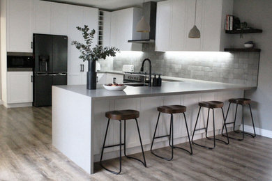 Inspiration for a mid-sized contemporary u-shaped open plan kitchen with an undermount sink, shaker cabinets, white cabinets, quartz benchtops, grey splashback, porcelain splashback, black appliances, laminate floors, with island, brown floor and grey benchtop.