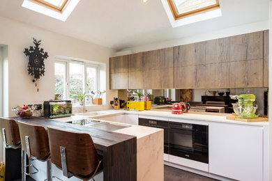 Small modern u-shaped open plan kitchen in London with an integrated sink, flat-panel cabinets, white cabinets, composite countertops, metallic splashback, mirror splashback, black appliances, ceramic flooring, a breakfast bar and brown floors.