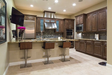 Example of a mid-sized transitional l-shaped travertine floor eat-in kitchen design in Las Vegas with a double-bowl sink, shaker cabinets, brown cabinets, granite countertops, gray backsplash, glass tile backsplash, stainless steel appliances and an island