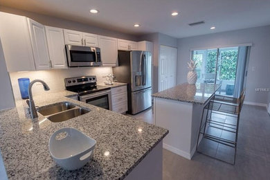 Open concept kitchen - mid-sized coastal l-shaped porcelain tile and gray floor open concept kitchen idea in Tampa with a double-bowl sink, raised-panel cabinets, white cabinets, granite countertops, white backsplash, ceramic backsplash, stainless steel appliances and an island