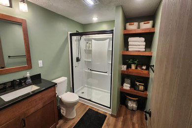 Mid-sized transitional 3/4 vinyl floor, brown floor and single-sink bathroom photo in Other with shaker cabinets, medium tone wood cabinets, a two-piece toilet, green walls, an undermount sink, granite countertops, black countertops and a built-in vanity