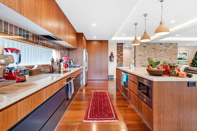 Design ideas for an expansive beach style galley open plan kitchen in Sydney with a double-bowl sink, beaded inset cabinets, medium wood cabinets, marble benchtops, multi-coloured splashback, brick splashback, stainless steel appliances, medium hardwood floors, with island, brown floor, white benchtop and recessed.