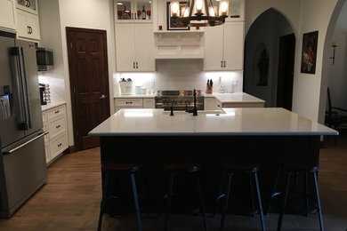 This is an example of a large arts and crafts l-shaped open plan kitchen in Orlando with a farmhouse sink, shaker cabinets, white cabinets, quartz benchtops, white splashback, subway tile splashback, stainless steel appliances, porcelain floors, with island, brown floor and white benchtop.