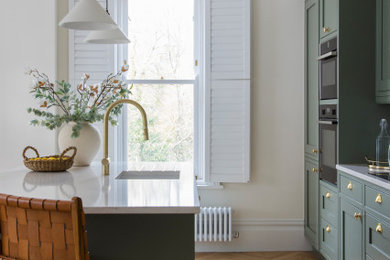 Design ideas for a small contemporary single-wall open plan kitchen in London with an integrated sink, shaker cabinets, green cabinets, quartz worktops, white splashback, ceramic splashback, integrated appliances, light hardwood flooring, an island and white worktops.