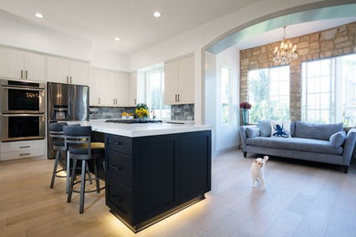 Inspiration for a large contemporary l-shaped open plan kitchen in San Diego with a farmhouse sink, shaker cabinets, black cabinets, quartz benchtops, grey splashback, marble splashback, stainless steel appliances, light hardwood floors, with island, beige floor and white benchtop.
