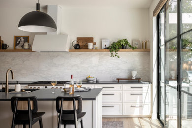 Large transitional l-shaped light wood floor open concept kitchen photo in Austin with an undermount sink, shaker cabinets, white cabinets, soapstone countertops, white backsplash, marble backsplash, white appliances, an island and black countertops