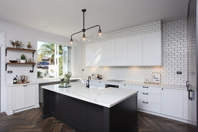 Photo of a large country u-shaped open plan kitchen in Sydney with a farmhouse sink, shaker cabinets, white cabinets, solid surface benchtops, white splashback, subway tile splashback, black appliances, porcelain floors, with island, brown floor and white benchtop.
