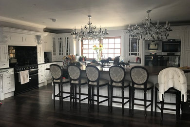 Photo of a large traditional open plan kitchen in Essex with a farmhouse sink, raised-panel cabinets, granite benchtops, black splashback, stone slab splashback, stainless steel appliances, dark hardwood floors, with island, brown floor and black benchtop.