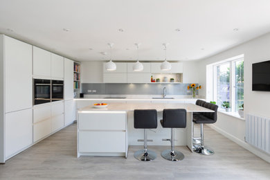 Photo of a medium sized contemporary l-shaped kitchen/diner in Gloucestershire with flat-panel cabinets, white cabinets, engineered stone countertops, grey splashback, glass sheet splashback, black appliances, an island and white worktops.