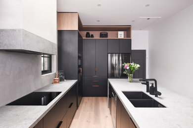 Photo of a large industrial l-shaped open plan kitchen in Sydney with an undermount sink, flat-panel cabinets, black cabinets, grey splashback, black appliances, light hardwood floors, with island, brown floor, grey benchtop and exposed beam.