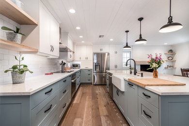 This is an example of a mid-sized country l-shaped eat-in kitchen in Tampa with a farmhouse sink, shaker cabinets, white cabinets, quartz benchtops, white splashback, ceramic splashback, stainless steel appliances, medium hardwood floors and with island.
