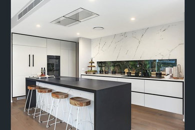 Photo of a large contemporary galley kitchen pantry in Brisbane with an undermount sink, flat-panel cabinets, white cabinets, quartz benchtops, window splashback, black appliances, with island, white splashback, light hardwood floors and brown floor.