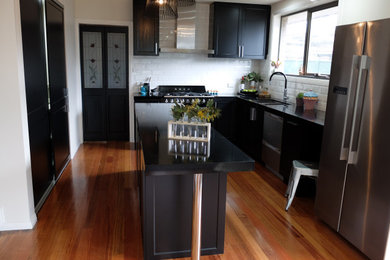 Mid-sized modern u-shaped kitchen pantry in Melbourne with a drop-in sink, flat-panel cabinets, black cabinets, white splashback, ceramic splashback, black appliances, with island, brown floor and black benchtop.
